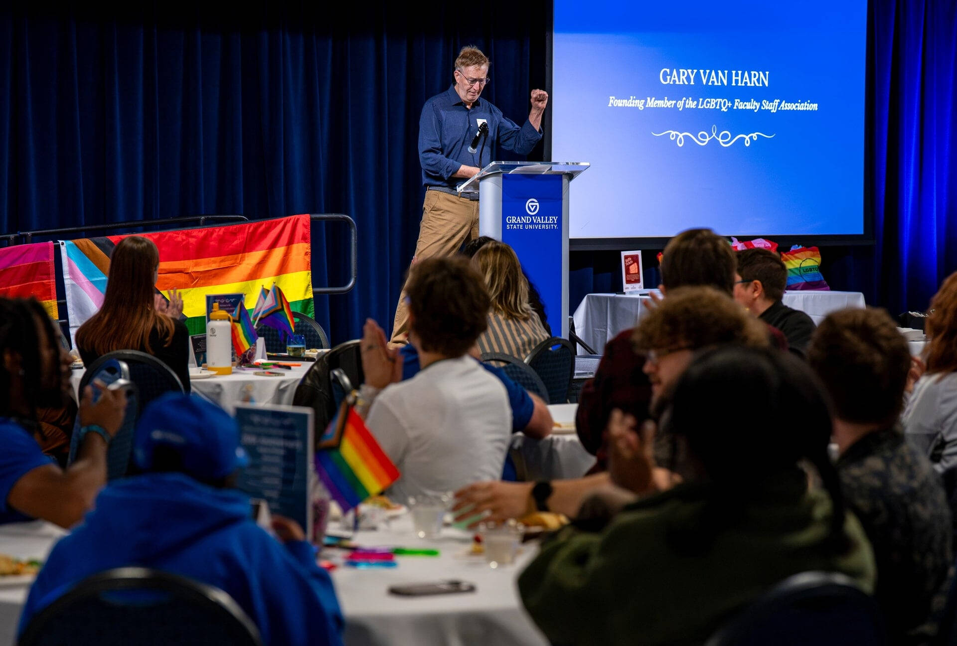 Gary Van Harn stands at podium to give remarks, people seated at tables in front; tables and stage decorated with rainbow flags